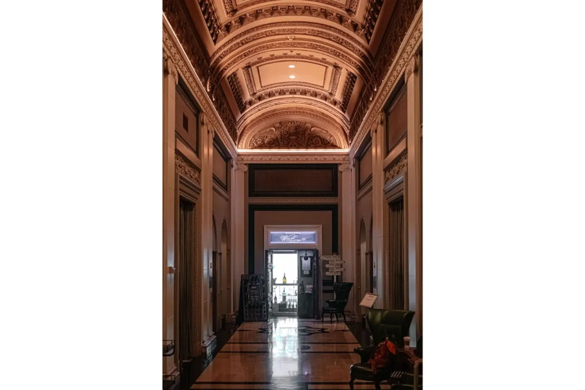 A wide hallway with an ornate ceiling, decorative moldings, and a doorway that leads outside. The space feels open and inviting, hinting at the transition between the sophisticated interior and the vibrant city beyond.