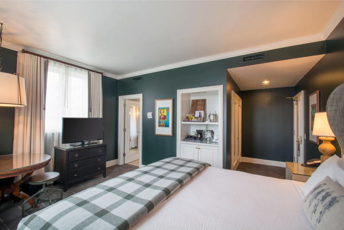 A welcoming bedroom scene featuring a comfortable bed with a plaid throw, a wooden table and chairs perfect for work or leisure, a TV atop a dresser, and vibrant artwork on the wall. Natural light fills the space, showcasing a blend of comfort and artistic flair.