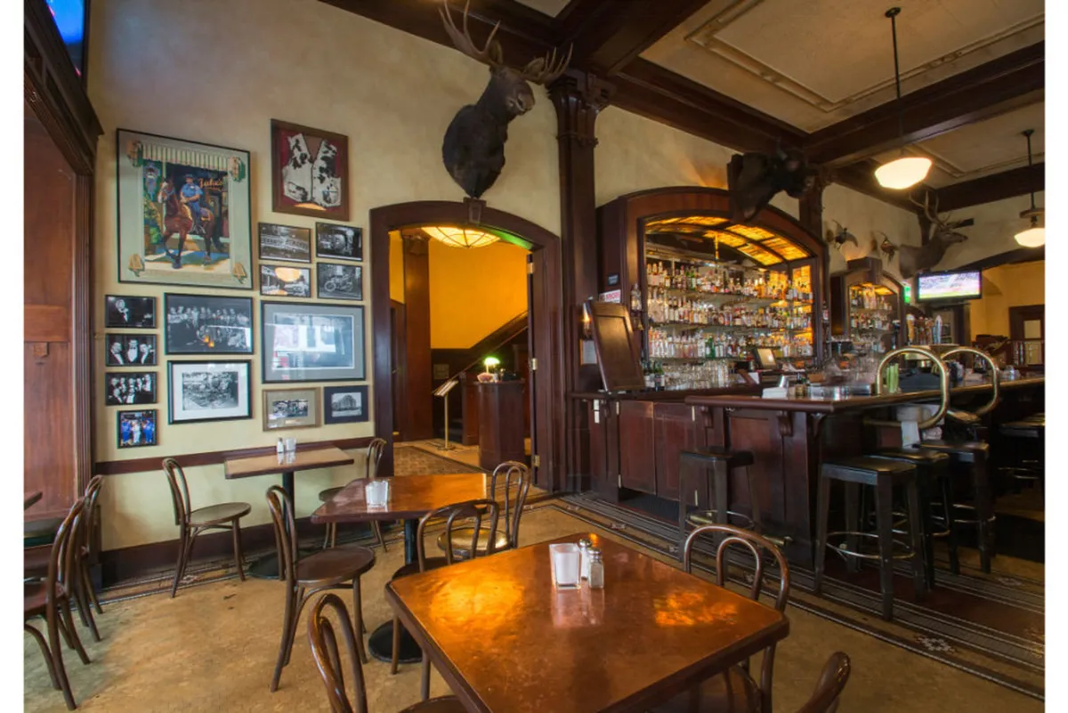 Inside, this cozy bar scene showcases a wide selection of bottles behind a wooden bar, with mounted animal heads and atmospheric lighting. Guests are seen socializing, enjoying drinks, and soaking in the relaxed, classic ambiance.