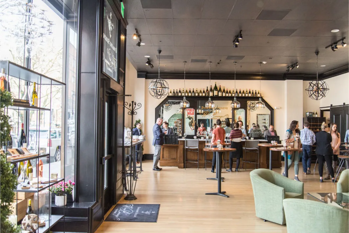 A lively bar area with a counter, hanging light fixtures, and an array of wine bottles on display. The space features ample seating for small gatherings or casual meetings, creating an inviting atmosphere for tasting events and social interactions.