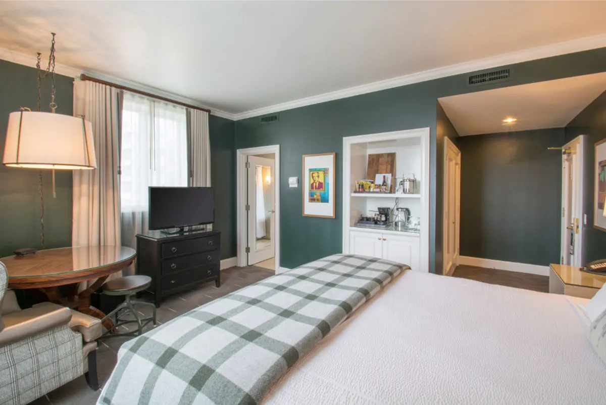 The image showcases a spacious and stylish hotel suite in Portland, Oregon. A plush king-sized bed with a patterned throw blanket serves as a cozy focal point. The room features a sleek TV unit, a round dining table perfect for intimate meals or morning coffee, and curated artwork that adds a sophisticated touch. The setting exudes comfort and warmth, inviting guests to relax, unwind, and enjoy both restful nights and vibrant city adventures.