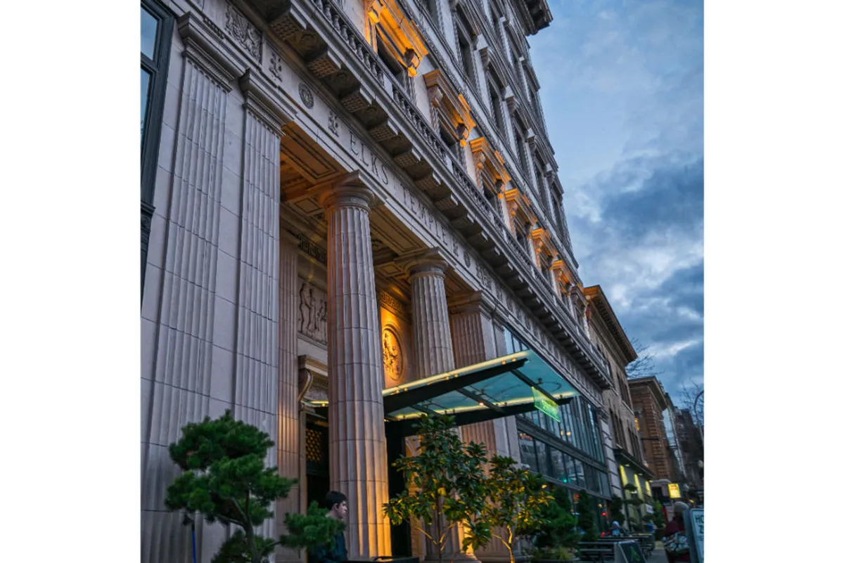 A wide exterior shot featuring grand columns, an illuminated entrance, decorative facade, and lively greenery in the foreground. The scene captures the energy of the surrounding street and the building’s sophisticated ambiance.