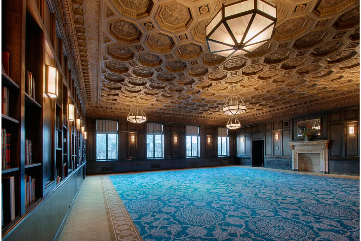 A refined, inviting lounge featuring an ornate ceiling, glittering chandeliers, a stately fireplace, blue patterned carpet, and walls lined with bookshelves. The space feels both grand and cozy, suited for literary discussions or enjoying art in a warm setting.