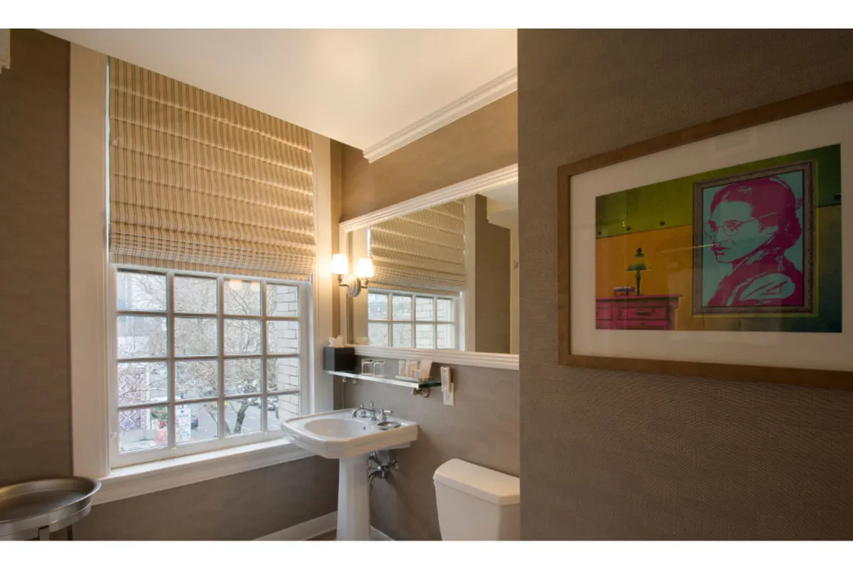 A modern bathroom scene with a sleek sink, framed artwork, and a window with blinds that allows morning light to filter in. The overall mood is calm and serene.