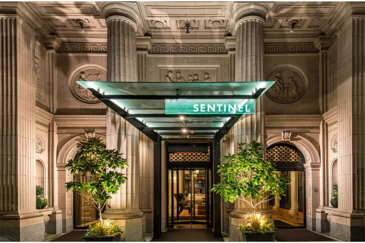 The exterior of the Sentinel hotel, highlighted by a glass canopy entrance, a prominent 'SENTINEL' sign, lush greenery, and classical architectural columns. The warm lighting and greenery create an inviting, sophisticated first impression.