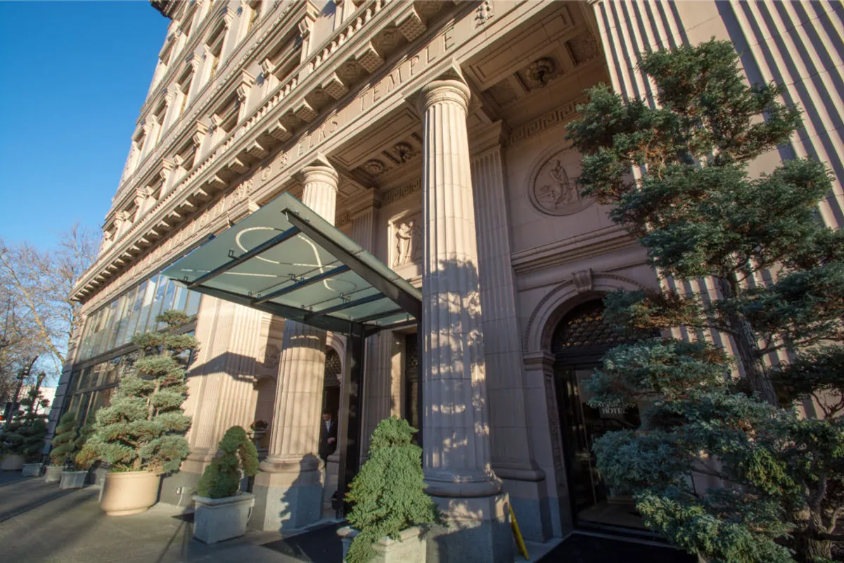 This exterior image features the hotel's grand entrance, complete with ornate columns, a glass canopy, and manicured shrubs. The setting conveys an inviting sense of arrival and hints at the luxury within.