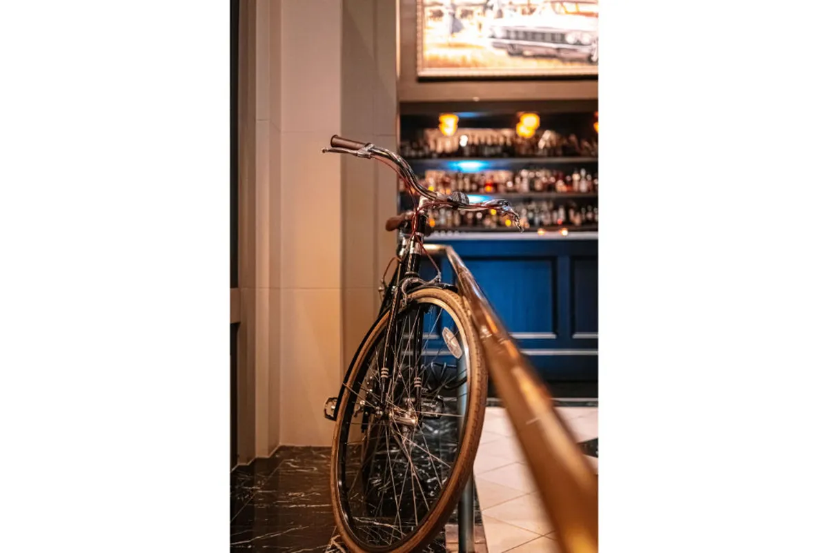 A stylish interior setting features a bicycle prominently displayed near a bar area with warm lighting fixtures, blending elements of urban culture and hospitality. The decor hints at the hotel's connection to city exploration and guest amenities.