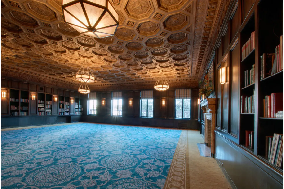 A serene, sophisticated library-like lounge featuring an ornate ceiling, large windows that let in abundant natural light, bookshelves lining the walls, intricate carpeting, and elegant chandeliers overhead. The space exudes calm and inspiration, perfect for reading or quiet work.