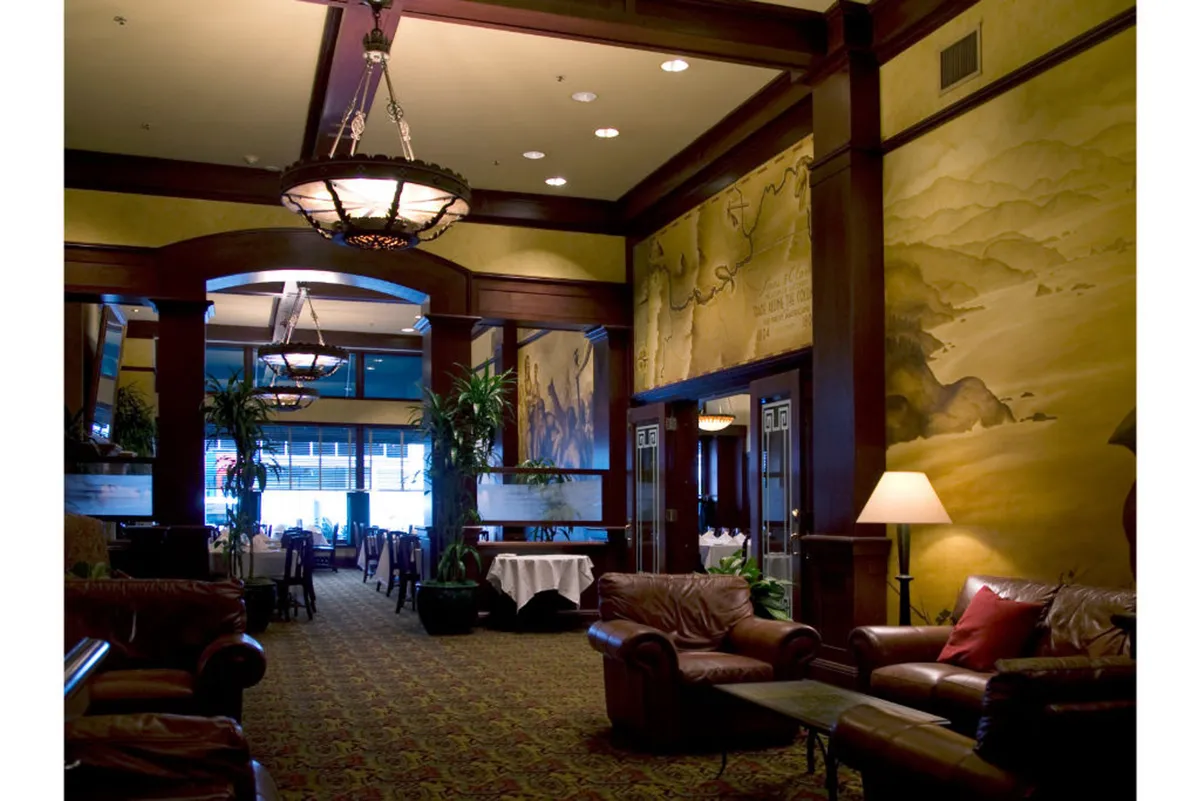 A sophisticated lounge space featuring bespoke Boys Fort furnishings, plush leather sofas, elegant chandeliers, and a decorative mural. The area is dotted with plants and a dining section where guests are engaged in relaxed social gatherings, dining, or quiet conversation.